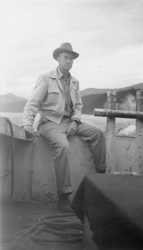 A man wearing a hat and a jacket sits on the side of a boat. There is a coiled rope on the floor and a view of mountains and water in the background.