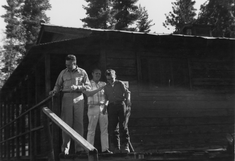 Three men standing on a wooden cabin porch. One is leaning on the railing, while another is holding a paper and standing next to him. The third man appears to be stepping down the stairs. Tall trees are visible in the background.
