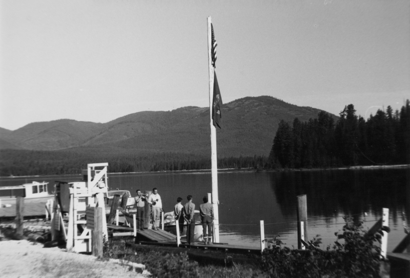 Several people stand on a dock beside a lake. A boat is moored nearby. Two flags are raised on a flagpole. In the background, there are trees and a range of hills or mountains.