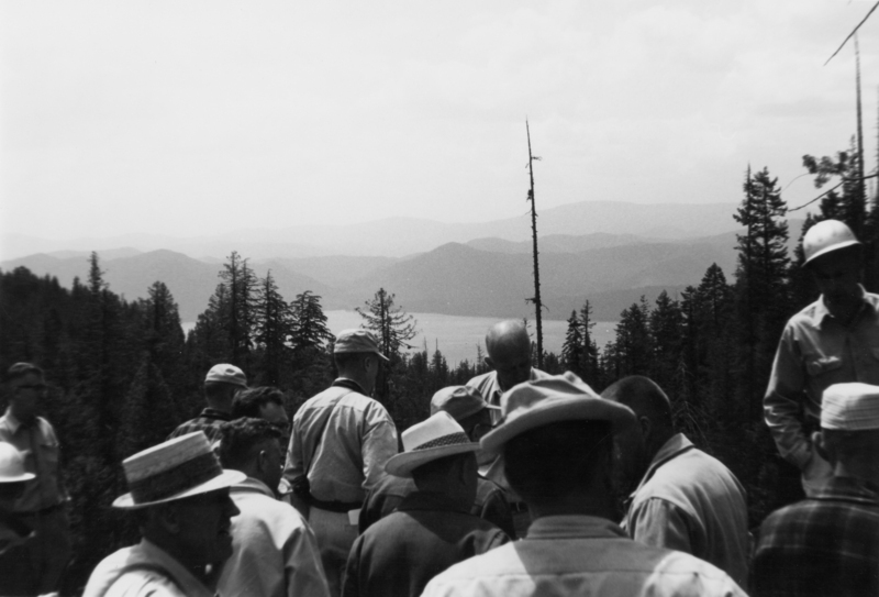 A group of people gathered in a forested area with mountains and a lake in the background. Many of the individuals are wearing hats and appear to be engaged in conversation.