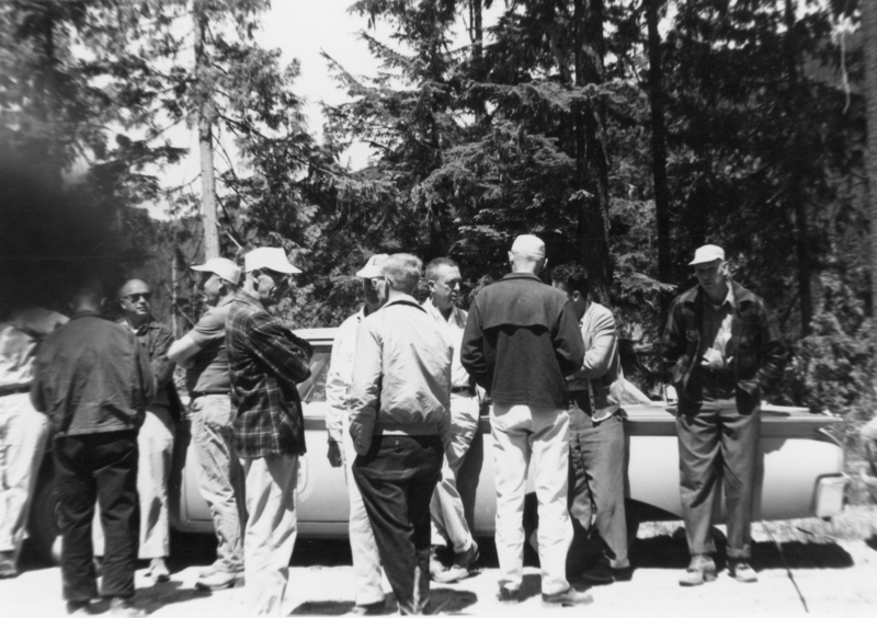 A group of men standing together outdoors near a car, surrounded by trees. Some are wearing hats and jackets.