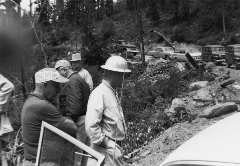 Several men wearing hats and jackets stand near a pile of debris in a forested area. There are cars parked on a dirt road in the background, and trees surround the scene.