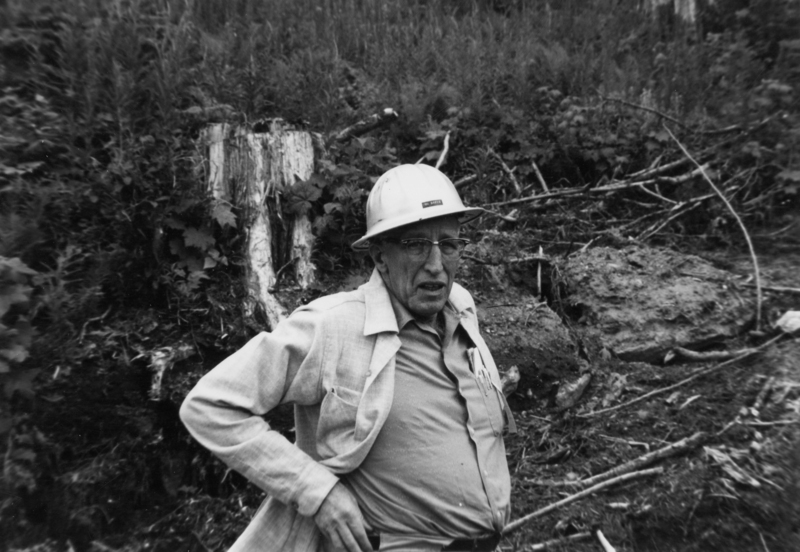 A man wearing a hard hat and glasses stands in a wooded area with uneven ground and scattered branches. He has a pen in his shirt pocket and is looking towards the camera.