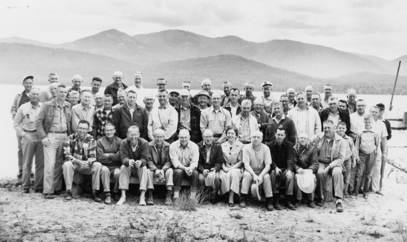 A large group of people, mostly men, posing together on a sandy area near a body of water, with mountains in the background. Some individuals are sitting in the front row while others are standing behind them. One person is wearing a hat with a letter on it, and there is a child among the adults.