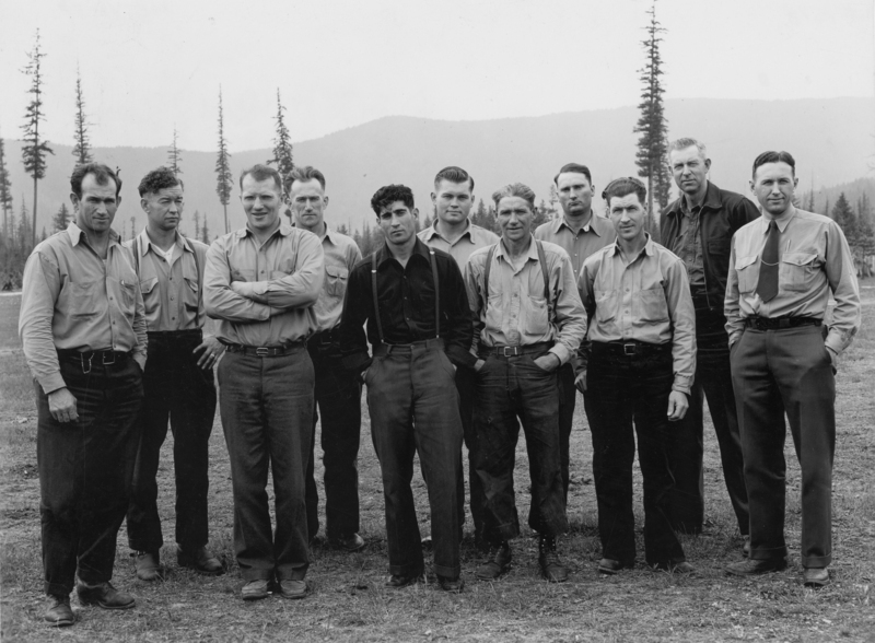 A group of eleven men standing together outdoors, wearing similar work shirts and pants, in a grassy area with trees and mountains in the background.
