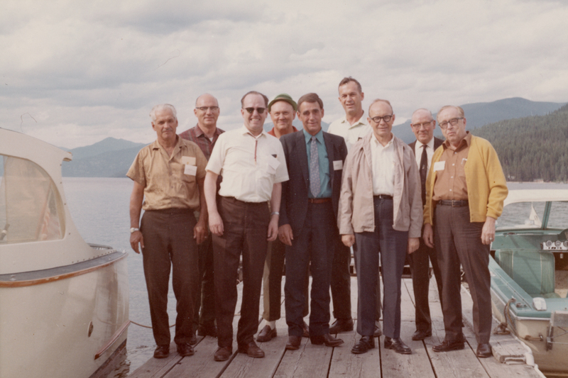 Nine men standing on a wooden dock near two boats. There is a large body of water and forested hills in the background. Some men are wearing eyeglasses, and they are dressed in a mix of casual and formal attire. They appear to be smiling at the camera.