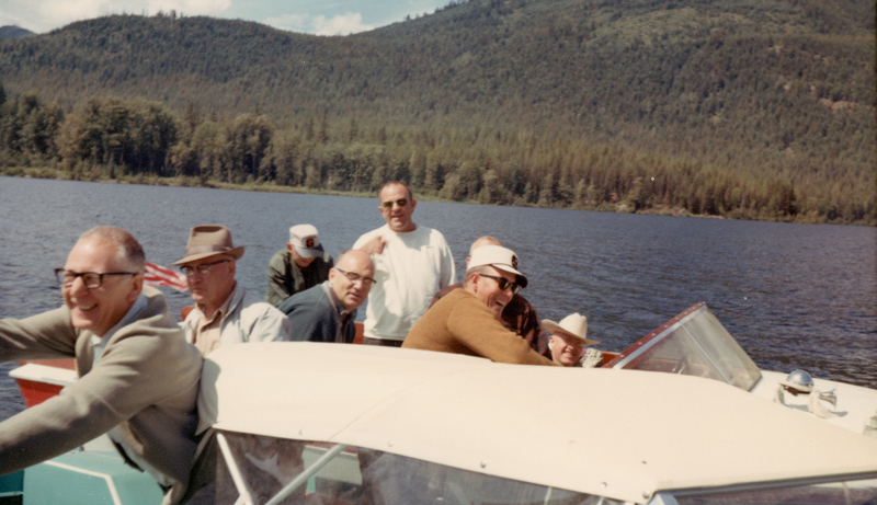 A group of men is gathered in a boat on a lake, with a forested hillside in the background. Some of the men are wearing hats and sunglasses, and they appear to be enjoying their time on the water.