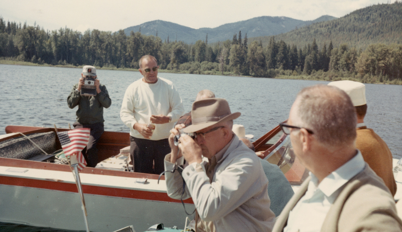 A group of people on a boat, surrounded by a lake and forested mountains. One person is taking a photo, another is holding a Polaroid camera, and a third person has a drink. An American flag is visible on the boat.