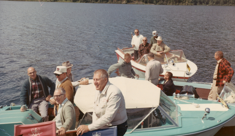 Several men are gathered on two motorboats on a lake. They are wearing casual outdoor clothing, including jackets, hats, and shirts. Some are sitting, while others are standing or leaning on the boats. The water is calm, and trees can be seen in the distance.
