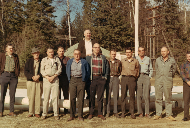 A group of twelve men standing outdoors in front of a wooded area and large poles. One man stands slightly behind the others. Most are wearing casual attire like shirts, sweaters, and jackets. The background includes trees and part of a structure.