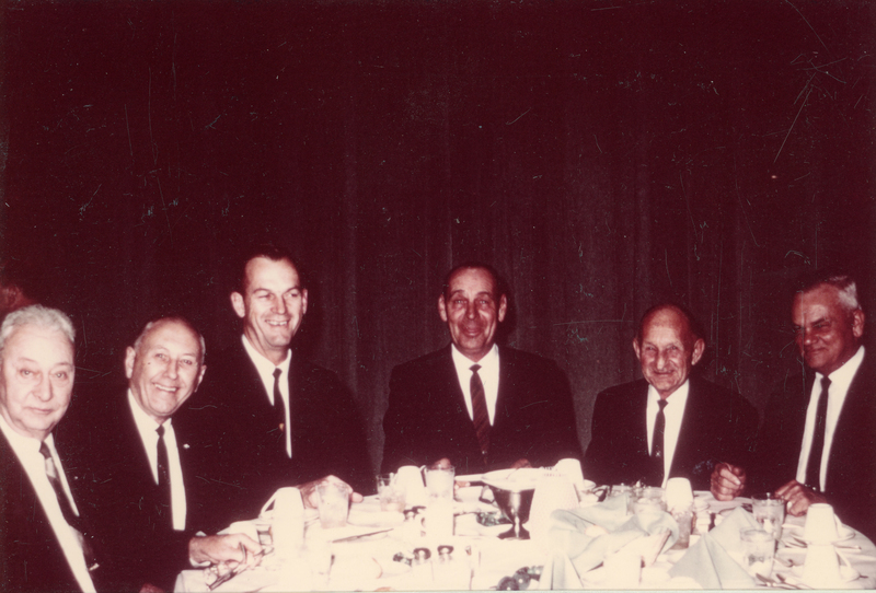 Six men in suits sitting at a table with tableware and drinks, appearing to enjoy a gathering or event.