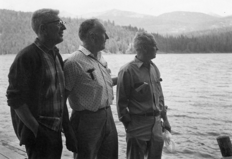 Three men stand together on a wooden deck with a large body of water and forested hills in the background. Each man has one hand in their pocket, and two of them are wearing glasses. One man holds a small object in his hand.