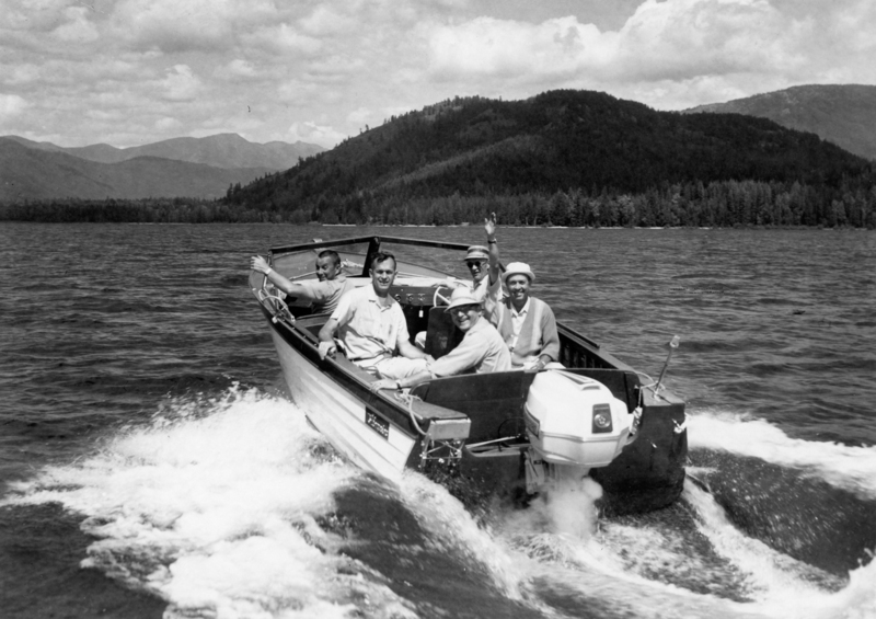 A group of five men is riding in a small boat on a lake. They are smiling and waving as the boat moves through the water. Behind them, there is a scenic view of hills and trees.