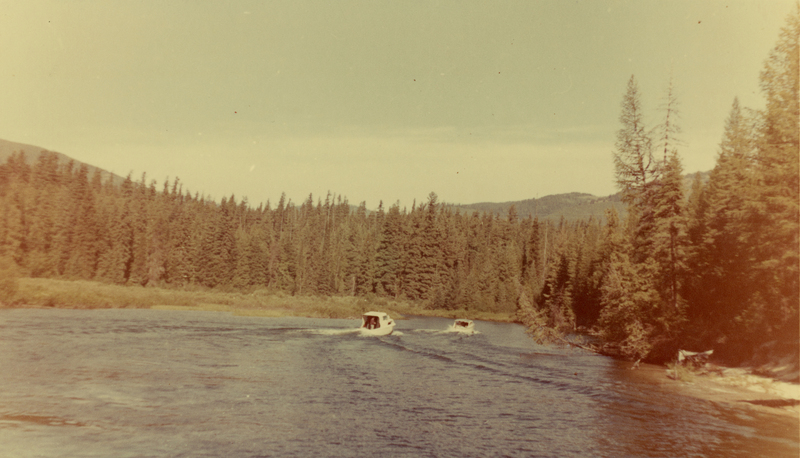 Two small boats are moving along a river, surrounded by a dense forest of tall trees with a mountain in the background.