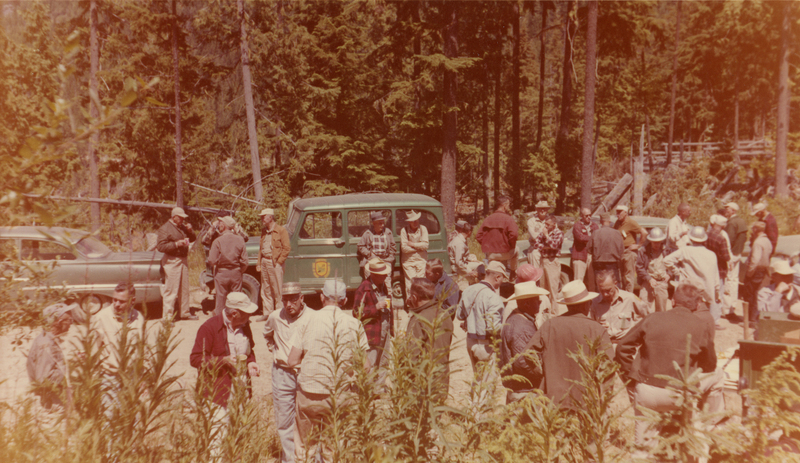 A large group of people gathered outdoors in a forested area with tall trees. Several individuals are engaged in conversation near parked vehicles. Some people are wearing hats and casual clothing, and greenery is visible in the foreground.