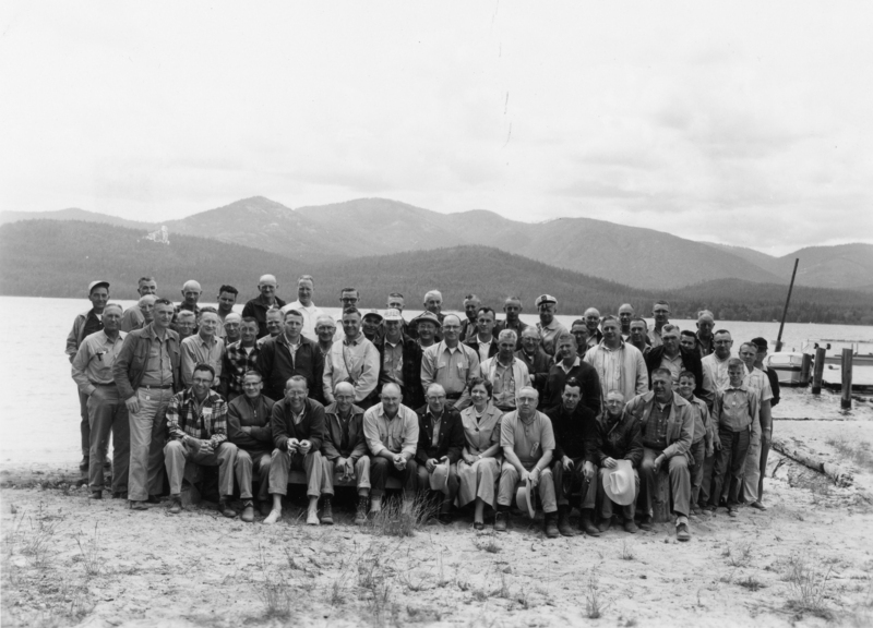 A large group of people, mostly men, gathered on a sandy beach with mountains and water in the background. Some are sitting in front, and others are standing behind. Several are wearing hats and casual outdoor clothing. A dock and a few boats can be seen in the background to the right.