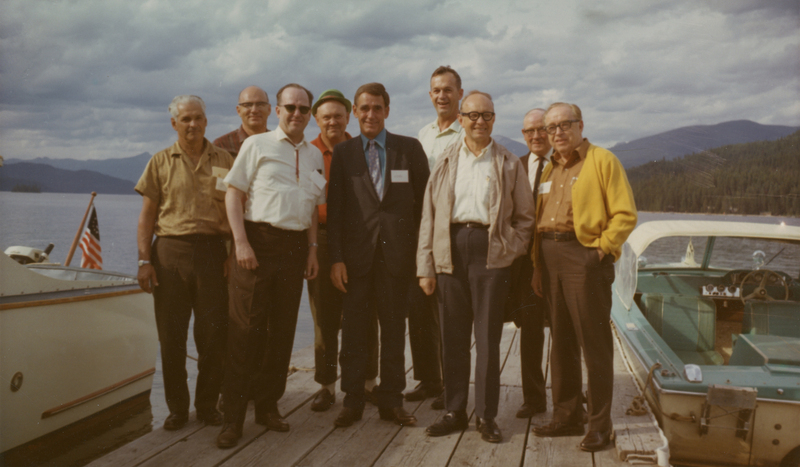 A group of nine men standing together on a wooden dock by a lake, with boats visible on either side and mountains in the background.