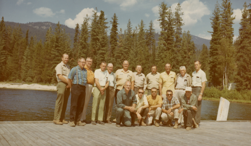 A group of sixteen men standing on a wooden dock near a body of water with a forest and mountains in the background. Some men are standing, while a few are crouching in front. They are dressed in casual and work attire.