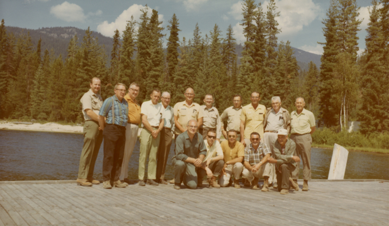 A group of men, some standing and some kneeling, on a wooden dock by a river with a forest and mountains in the background.