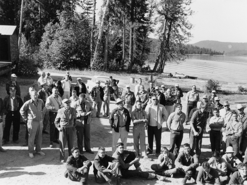 A group of men standing and sitting on a dirt area near a lake, surrounded by trees. Several men are wearing hats and jackets. A car is parked in the background. The scene includes a mix of standing individuals and others seated on the ground.