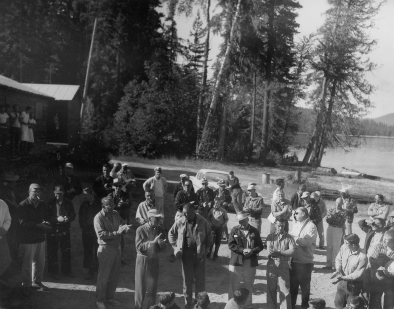 A group of people gathered outdoors near a building and surrounded by tall trees. Some are clapping, while others hold cups. In the background, a body of water and more trees are visible. Several people stand near a parked car, and a few are sitting or standing on a porch.