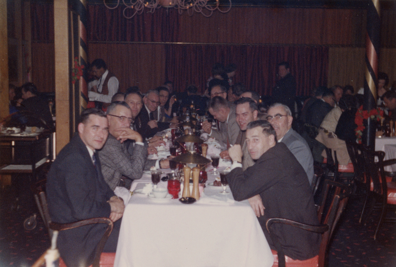 A group of men sitting around a long dining table set with dishes and glasses in a restaurant. The scene includes several men looking towards the camera with others engaged in conversation. The room has decorative elements, such as red curtains and patterned columns, and there are servers in the background.