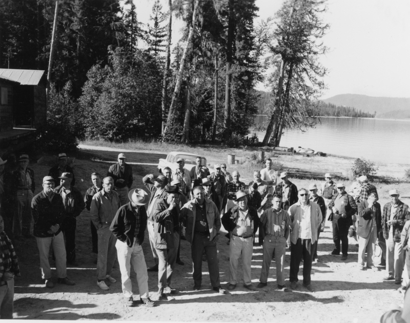 A group of people gathered outdoors near a building and trees, with a lake visible in the background. Some individuals are wearing hats, and most are looking in the same direction.