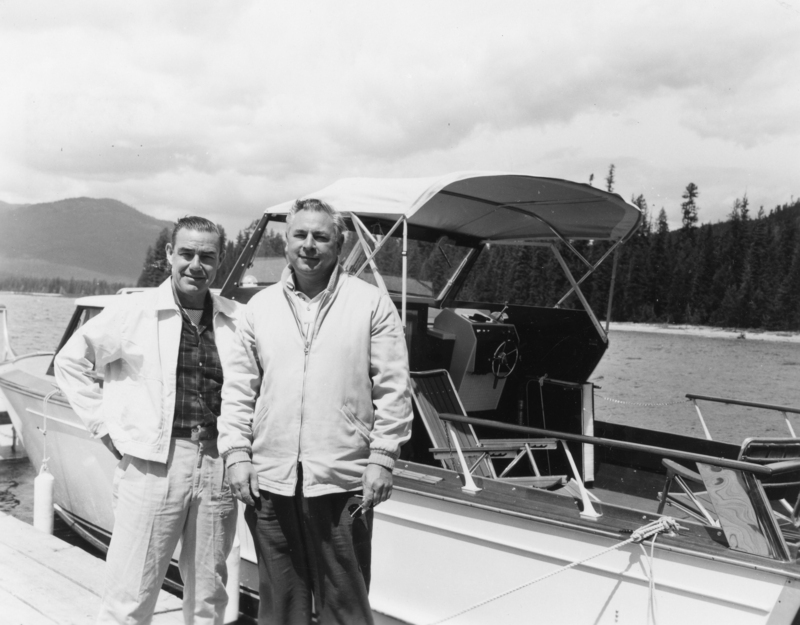 Two men standing on a dock beside a boat with a canopy. They are both wearing jackets. Behind them is a body of water with forested hills in the background.
