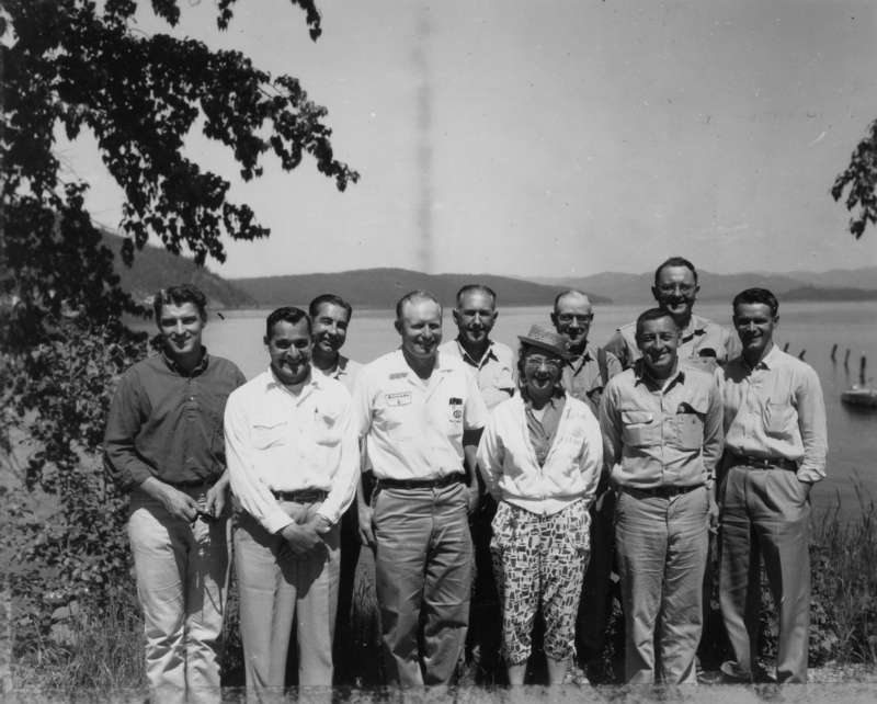 A group of nine people standing together near a body of water, with trees on either side and distant mountains in the background.
