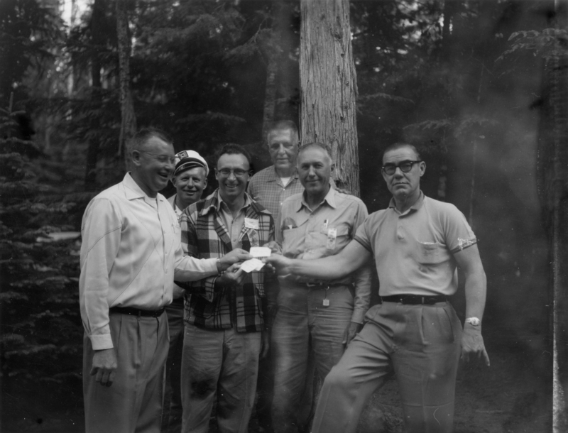 A group of six men standing together in a forest. One man is wearing a cap, and others are dressed in shirts and trousers. They are holding what appears to be a small piece of paper or card. The background is wooded with trees.