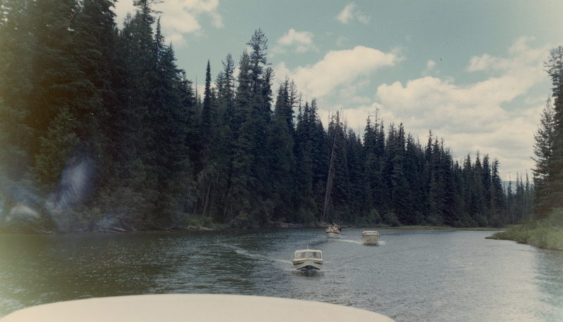 Boats moving along a river surrounded by dense forest with tall trees. The sky is partly cloudy.