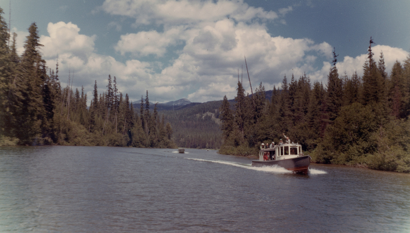 A small boat with several people on board traveling down a river surrounded by dense trees. A mountain range is visible in the background under a partly cloudy sky.