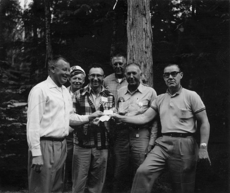 Six men standing together in a forested area, one holding a paper. They are dressed casually and appear to be enjoying a moment outdoors.