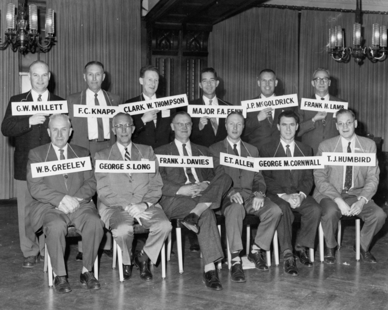 Group of men seated and standing in two rows, each holding name plaques. They are dressed in suits and ties and are positioned indoors with decorative light fixtures visible above them. Plaques read: "G.W. MILLETT" - "F.C. KNAPP" - "CLARK W. THOMPSON" - "MAJOR F.A. FENN" - "J.P. MCGOLDRICK" - "FRANK H. LAMB" - "W.B. GREELEY" - "GEORGE S. LONG" - "FRANK J. DAVIES" - "E.T. ALLEN" - "GEORGE M. CORNWALL" - "T.J. HUMBIRD".