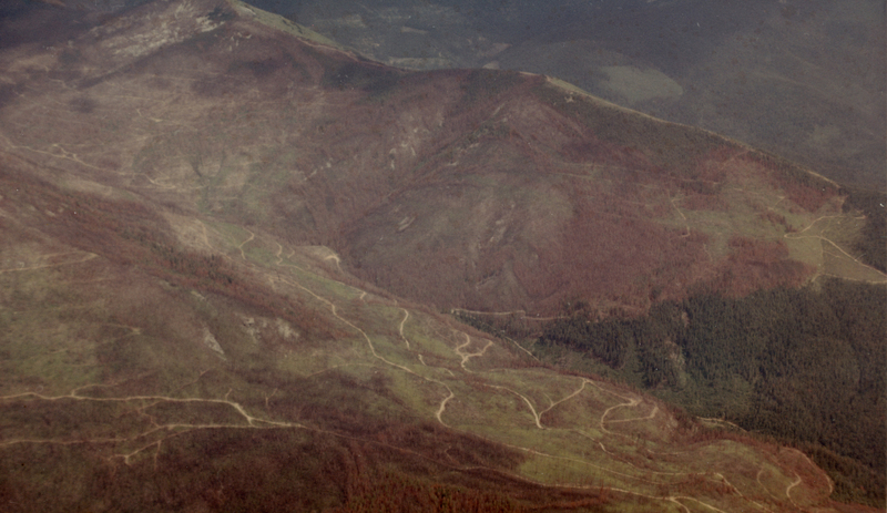An aerial view of a landscape with rolling hills and sparse vegetation. Winding paths are visible, crisscrossing the terrain. The hills have some areas covered with trees, while other parts appear open and barren. The scene stretches into the distance with overlapping ridges.