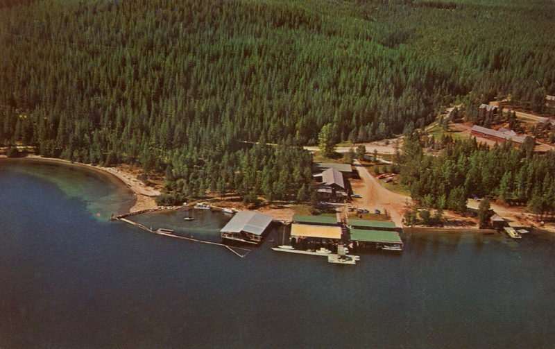 Aerial view of a forested area with a large body of water. There are buildings and a dock extending into the water, with several boats moored at the dock. A sandy shoreline curves along the edge of the water, and a road is visible among the trees.