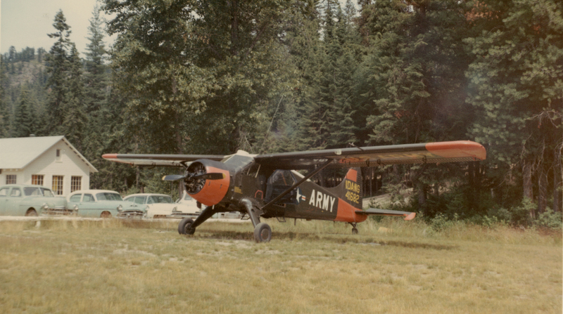 An airplane with markings on its side is parked on a grassy field. Behind it, there are several vintage cars and a building with a light-colored exterior. The area is surrounded by tall trees. National Guard plane at Cavanaugh Bay with text reading: "IDANG 41682" and "ARMY"