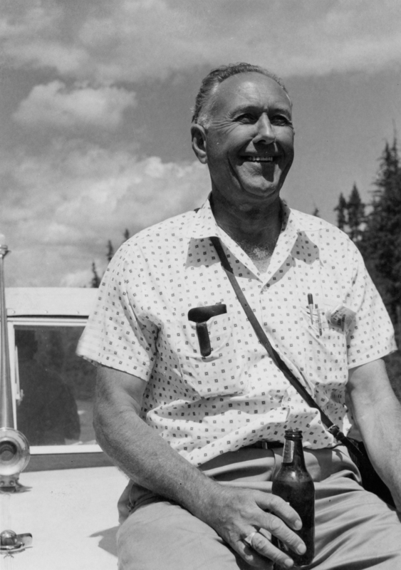A man with short hair is sitting outdoors holding a bottle. He is wearing a patterned short-sleeved shirt, with pens in the pocket and a strap across his chest. Behind him, a car is visible and there are trees and a cloudy sky in the background.