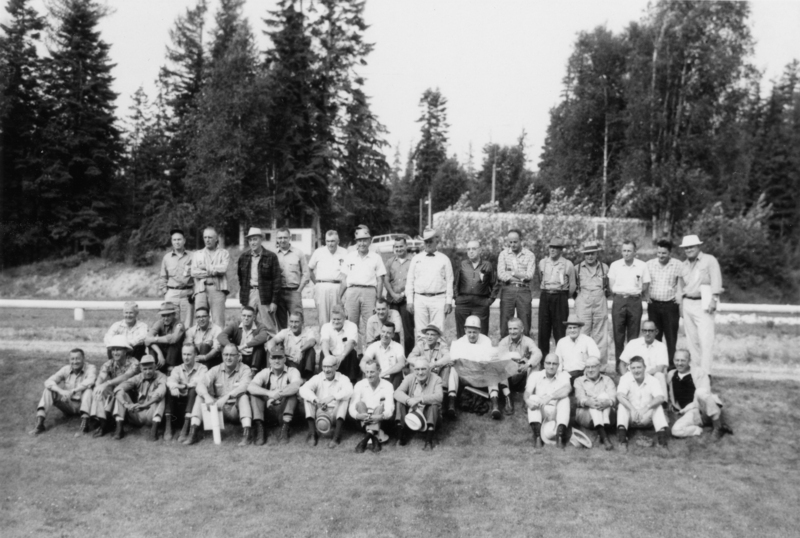 A group of men posed in front of a wooded area, with some sitting on the grass in front and others standing behind them. A few are wearing hats and several have their arms crossed. A pathway and trees are visible in the background.