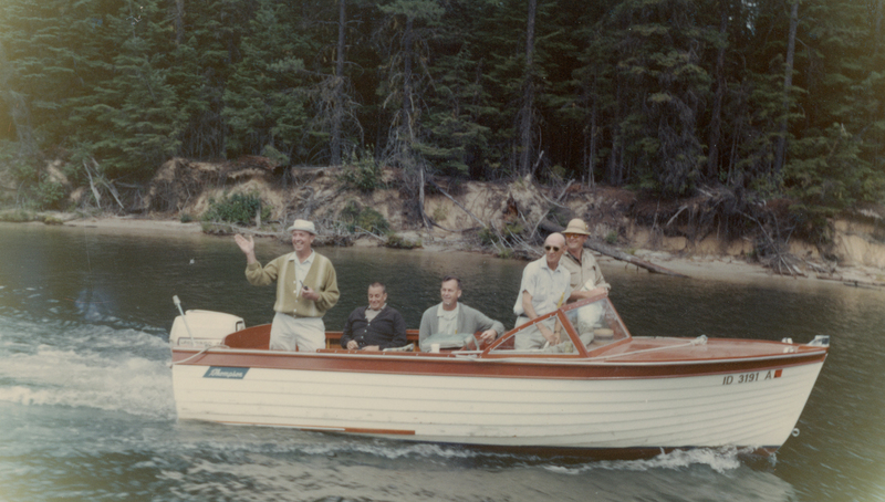 Five men are on a small motorboat traveling along a calm body of water, surrounded by lush forested terrain. One man, standing, gestures while holding an object, and the others sit or stand nearby. "Thompson" is written on the side of the boat and "ID 3191 A" near the front.