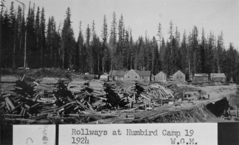 Stacks of logs are piled in the foreground, with several wooden buildings and a dense forest of tall trees in the background. Typed text near the bottom reads: "Rollways at Humbird Camp. 1924 W.G.M."