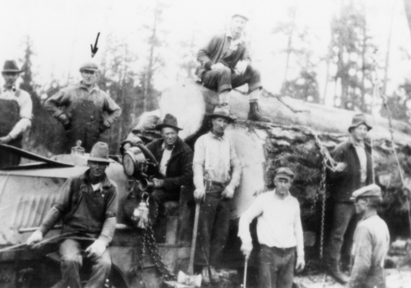A group of men gathered around a large log in a forest setting. One man sits on top of the log, while others stand or sit near a piece of machinery. They are dressed in work clothes and hats. One man has an ax, and another has chains near the log.
