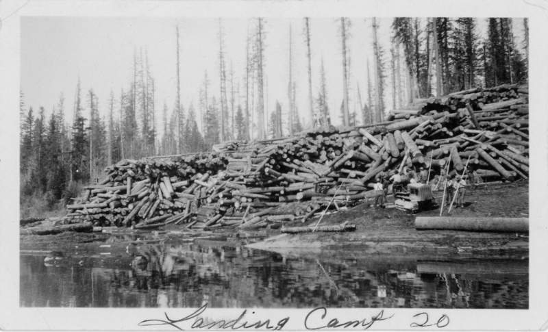 A large pile of logs stacked beside a body of water, with several people and a small piece of machinery in the foreground. Tall trees stand in the background. The handwritten text near the bottom reads "Landing Camp 20".