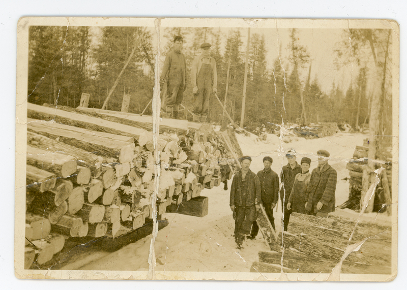 A group of men in winter clothing stand beside stacked logs in a snowy forest. Two of them are standing on top of the logs, holding long tools, while the others are positioned on the ground. Trees surround the scene in the background.