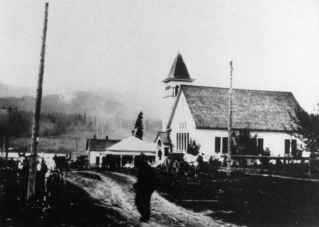 A figure is standing on a dirt road in front of a large building with a steep roof and a tower, possibly a church. There are horses with carriages parked along the road. Other small buildings are in the background, and trees can be seen in the distance.
