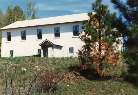 A rectangular building with a pointed roof and several windows, set in a grassy area with trees surrounding it.