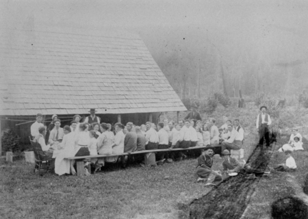 A large group of people is seated on benches around a long table outside near a wooden building with a sloped roof. Two men stand beside them, and several individuals are sitting on the grass nearby. The scene is set in a grassy area with trees in the background.