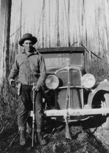 A man wearing a hat and jacket stands in front of an old car in a wooded area. He holds a walking stick, and a rifle is leaning against the car.