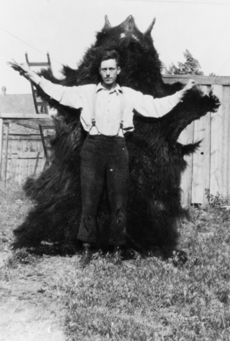 A man standing in a yard with his arms outstretched, holding a large bear hide behind him. There is a wooden fence and a ladder in the background.