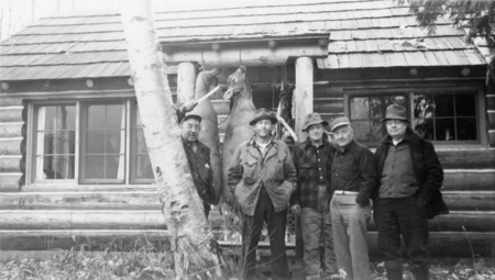 Five men standing in front of a log cabin, with two deer hanging behind them. A tree trunk is visible in the foreground. The men are dressed in outdoor clothing, including hats and jackets.